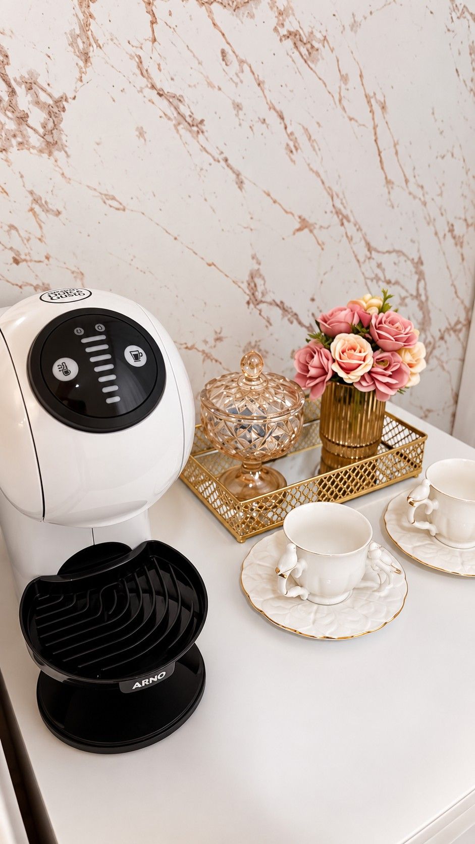 Cafeteira, bandeja dourada com flores e porta-balas de vidro, e duas xícaras de porcelana sobre a bancada branca, diante da parede marmorizada.