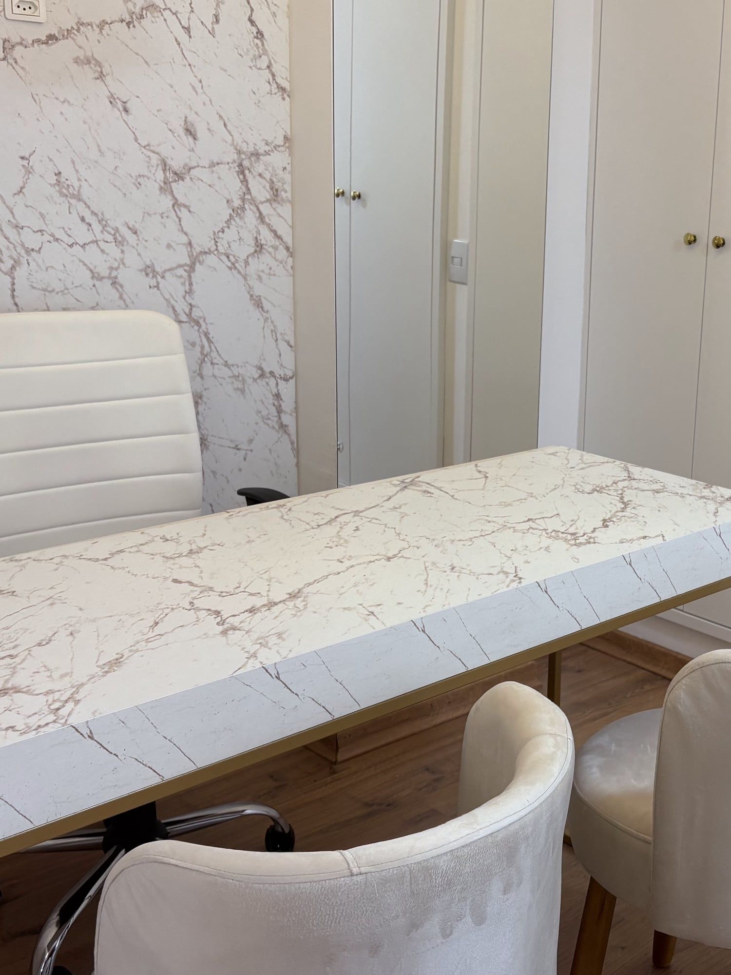 Mesa de atendimento em mármore branco com pés dourados, cadeira de escritório e duas poltronas de veludo, diante de armários brancos.
