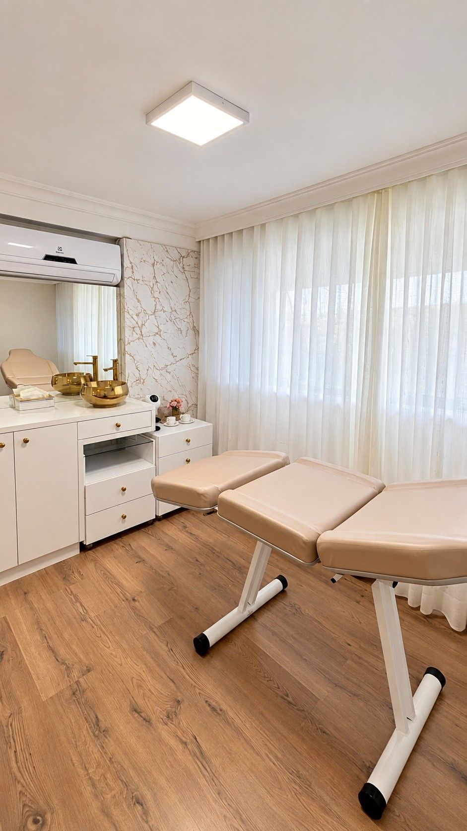 A sala inteira: maca reclinável bege em primeiro plano, bancada branca com cubas douradas e espelho à esquerda, ar-condicionado e cortina clara na janela.